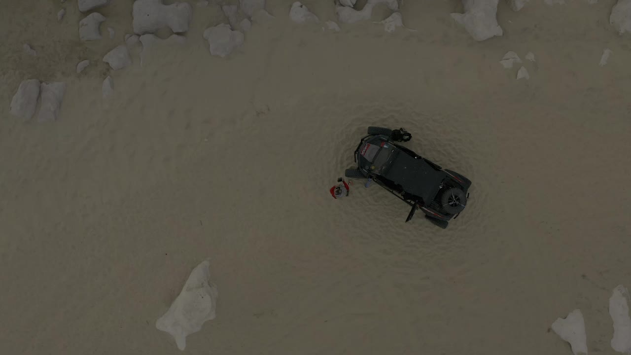 High overhead view in Baja California Sur, Mexico, with the dark vehicle angled on open sand, rocks clustered above it, and no recognizable people visible.