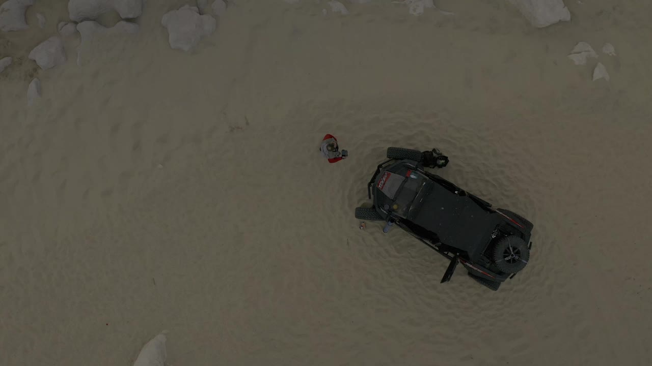 Child in red shirt and dark vehicle seen from directly overhead on sandy ground.