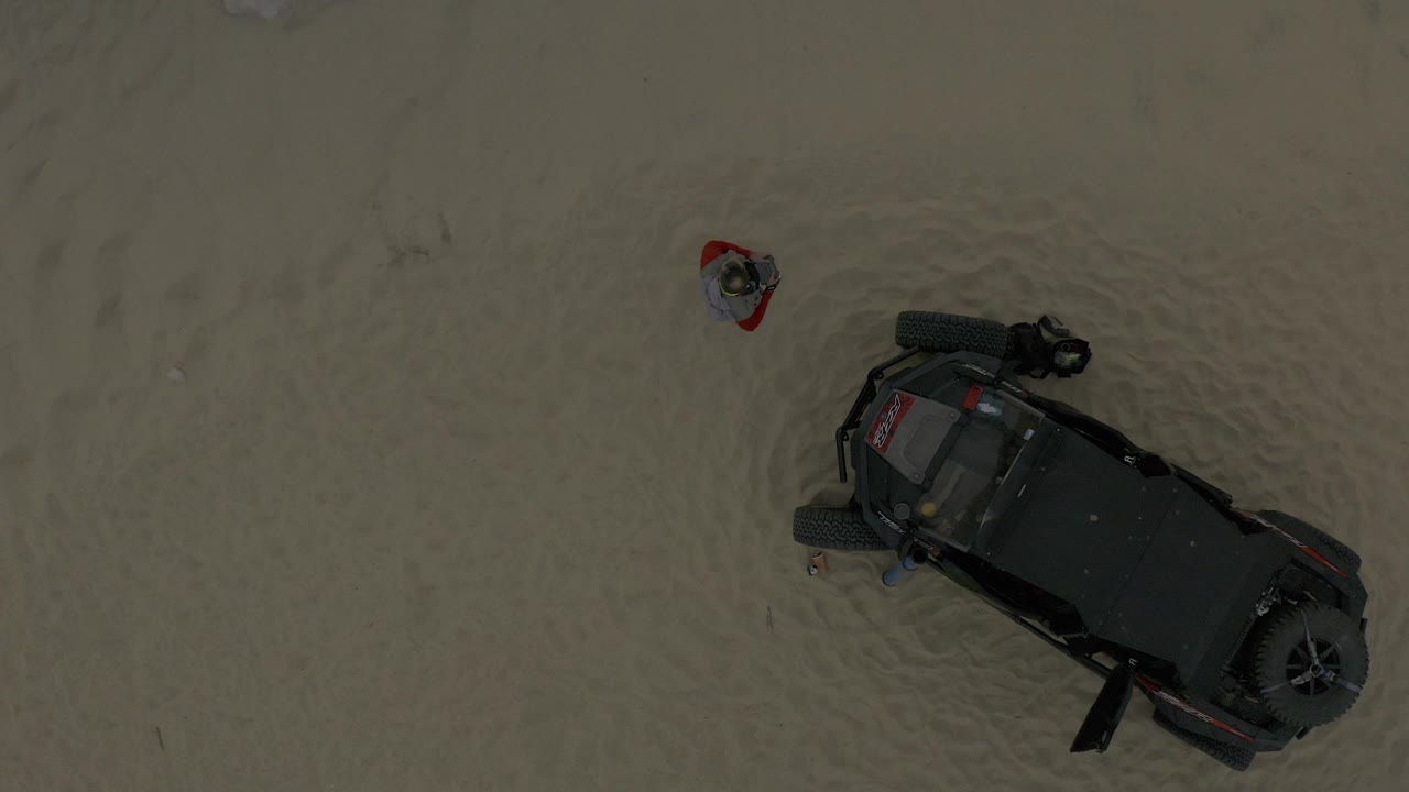 Overhead Baja California Sur Mexico view of a man in a red layer standing in pale shallow water near the upper side of a dark off-road vehicle.