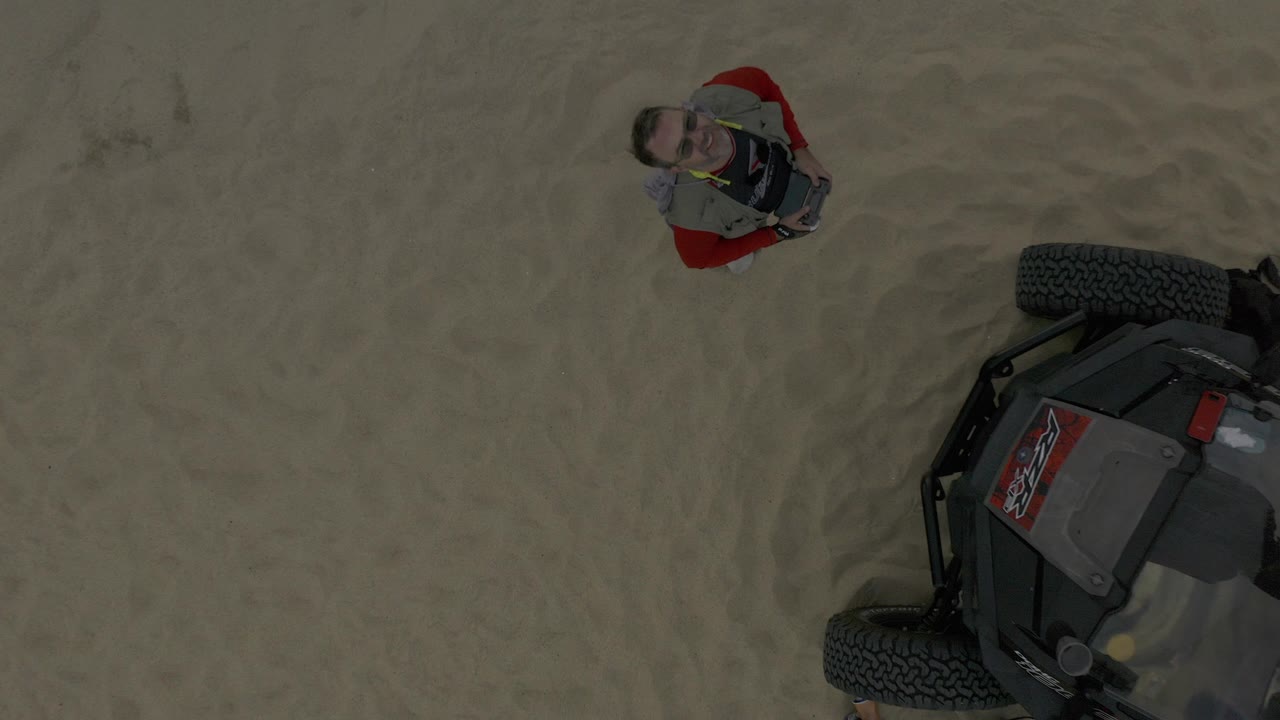 Overhead frame in Baja California Sur, Mexico showing a man in red clothing standing on rippled sand beside the cropped tire and body of an off-road vehicle.