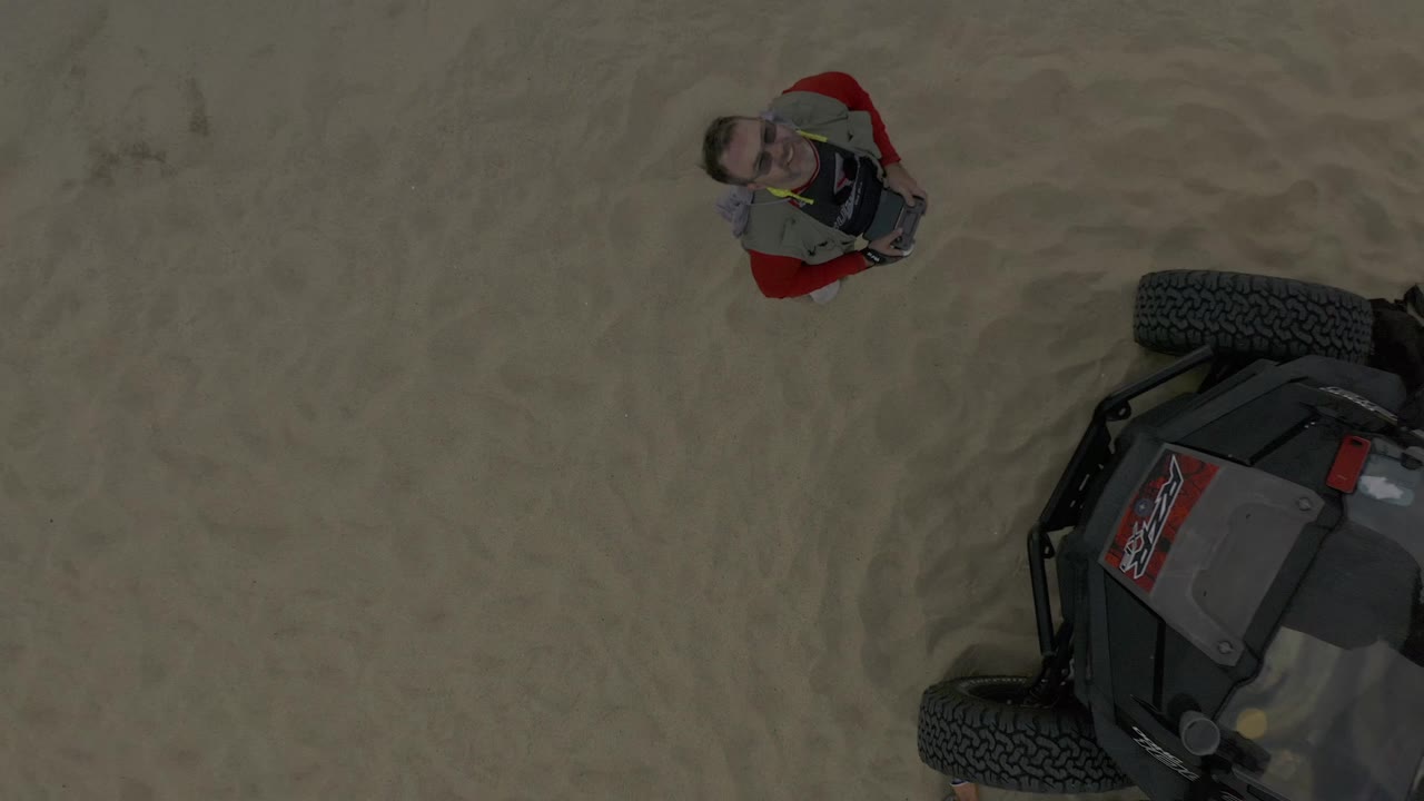 Man in red clothing stands on sand beside a cropped off-road vehicle.