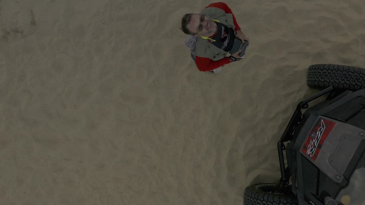 The Baja California Sur overhead composition shifts slightly, with the man in red higher in frame and the cropped off-road vehicle entering from the right edge over textured sand.