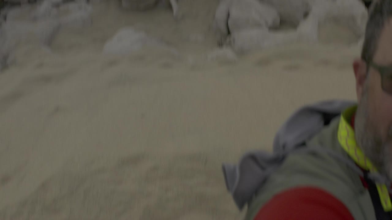 A partial person in a grey vest and red shirt appears at the right edge in Baja California Sur, Mexico, beside tan sand and pale shoreline rocks.
