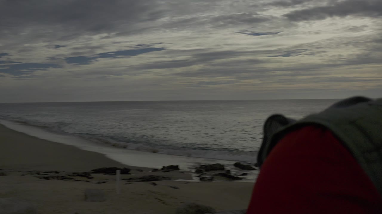 Baja California Sur beach view with gray sky, ocean horizon, sand, low surf, and a blurred red sleeve or jacket edge blocking part of the foreground.