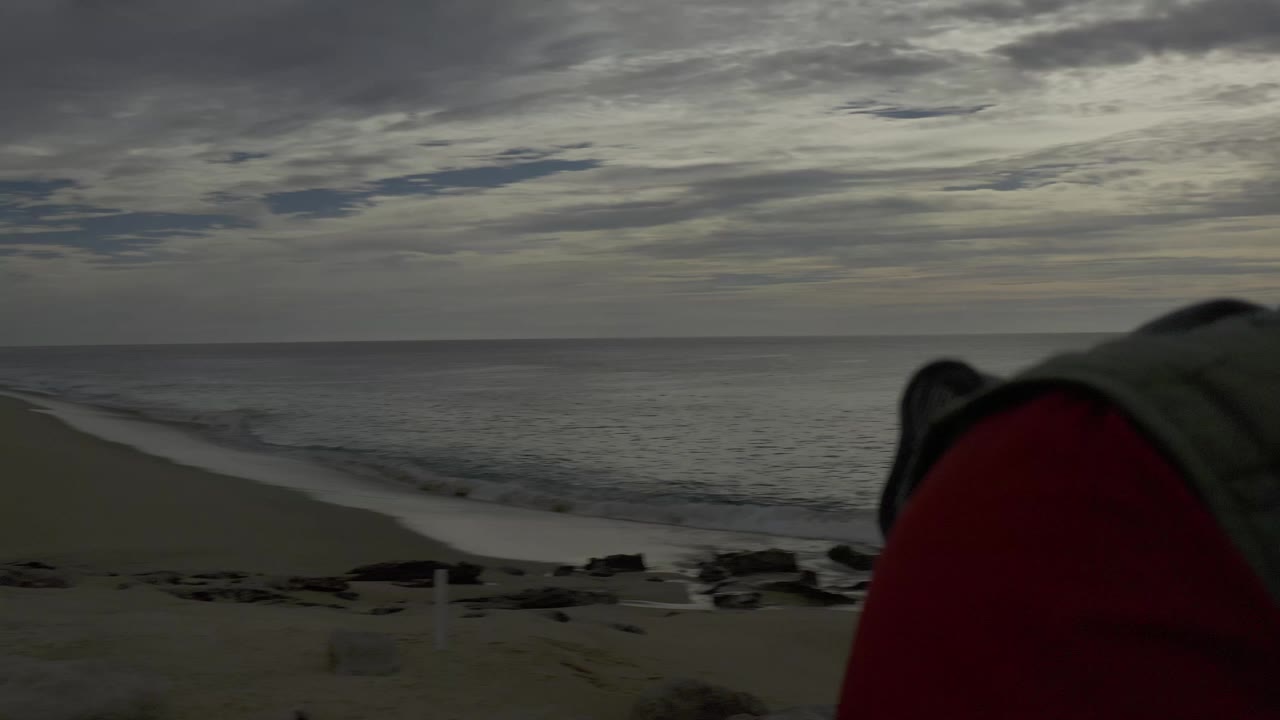 Baja California Sur shoreline under a cloudy sky, with the ocean horizon across the frame and a red and gray foreground shape intruding near the lower right.
