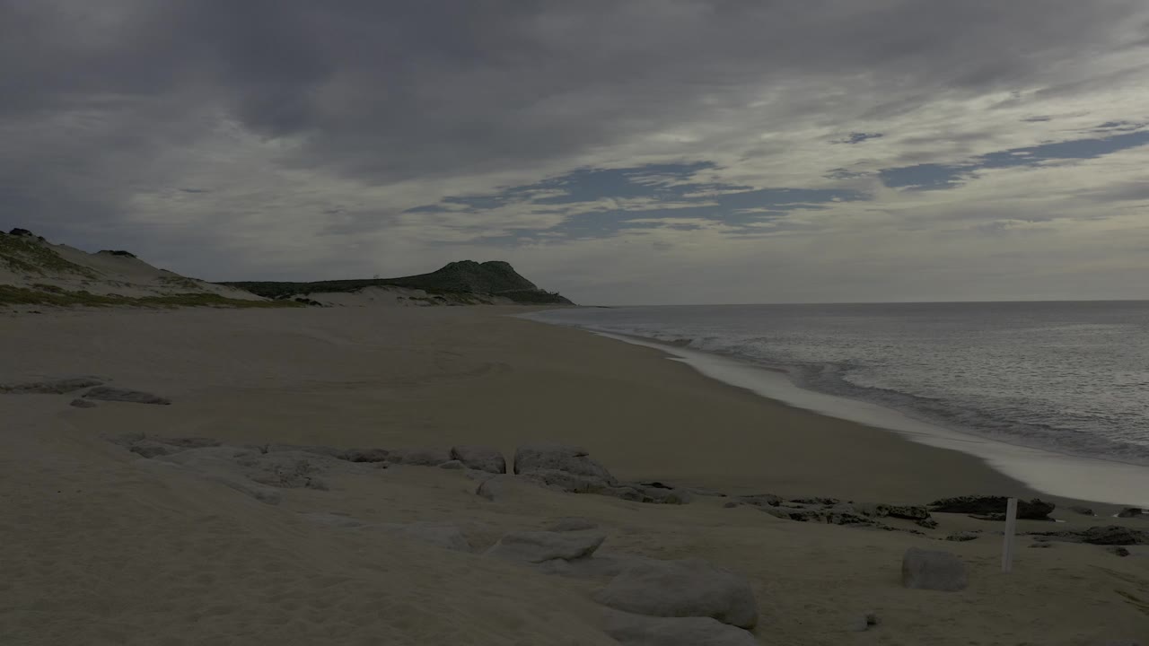 Wide Baja California Sur beach frame with sand in the foreground, low dunes or hills in the distance, ocean on both sides of the composition, and no visible people.