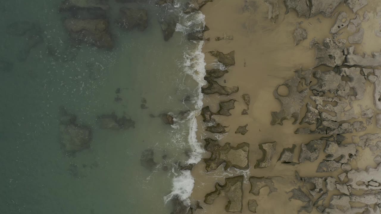 Overhead Baja California Sur beach view with the foam line slightly farther along the rocks, muted green water on the left, tan sand on the right, and no visible people.