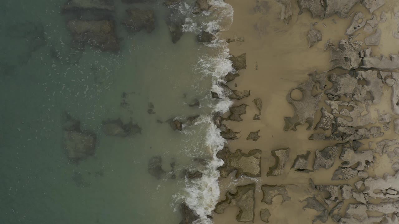 Straight-down view of the Baja California Sur shoreline as white surf spreads across dark rocks between green water and tan beach, with no visible people.