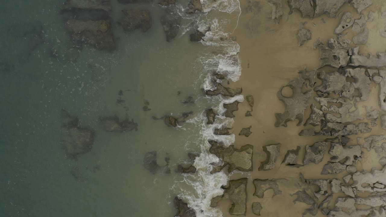 Top-down frame of Baja California Sur coast with more foam gathered around the central rock band, shallow green water to the left, sand to the right, and no visible people.