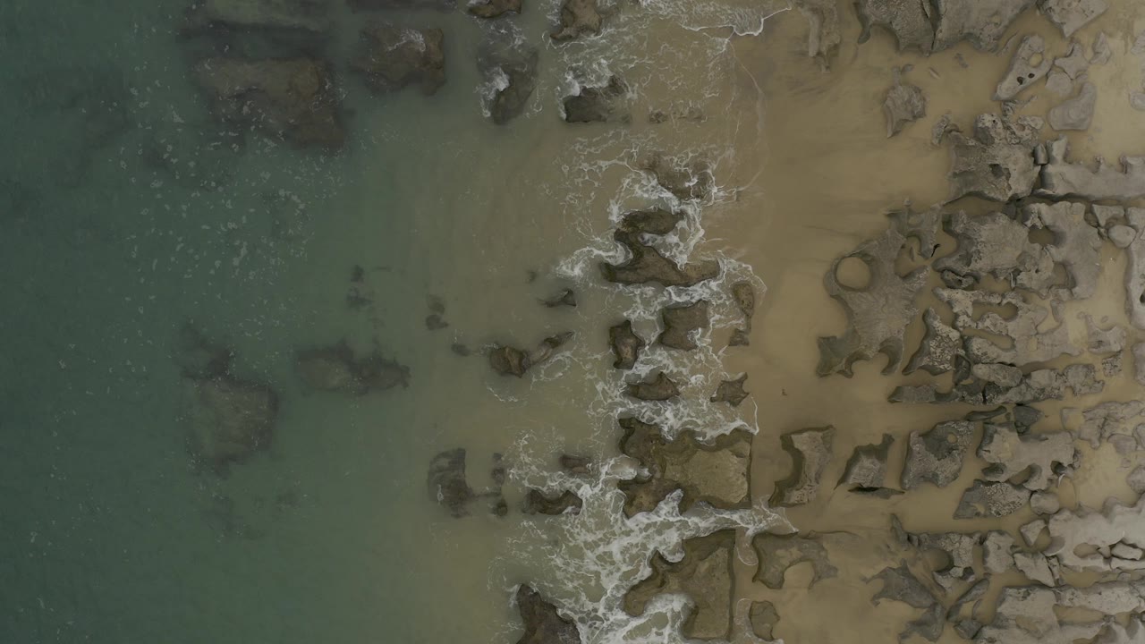 Aerial view of green water, tan sand, rocks, and light surf.