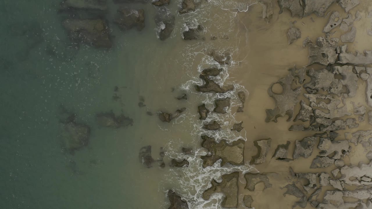 Aerial view in Baja California Sur, Mexico, where white foam curls around a central line of rocks between green water and tan sand, with no visible people.