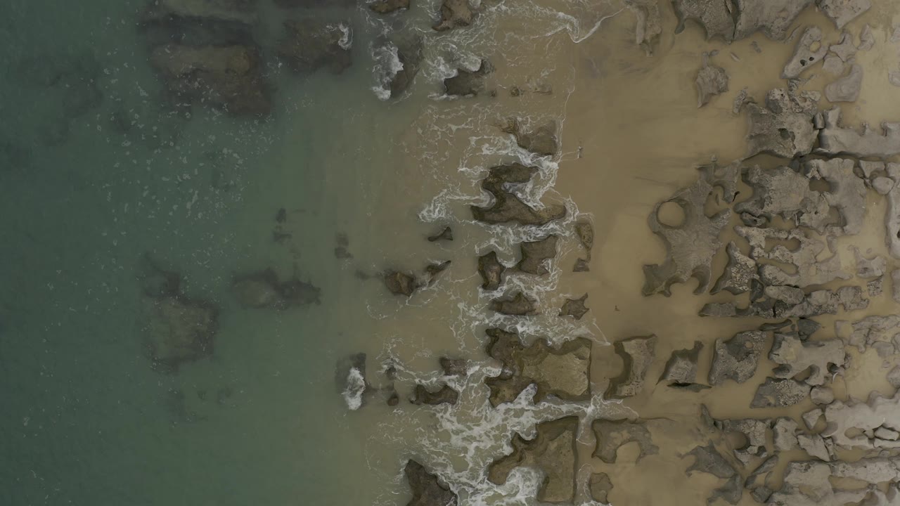 Top-down Baja California Sur shoreline frame with a small white breaker near the center-left, darker submerged rocks in green water, and no visible people.