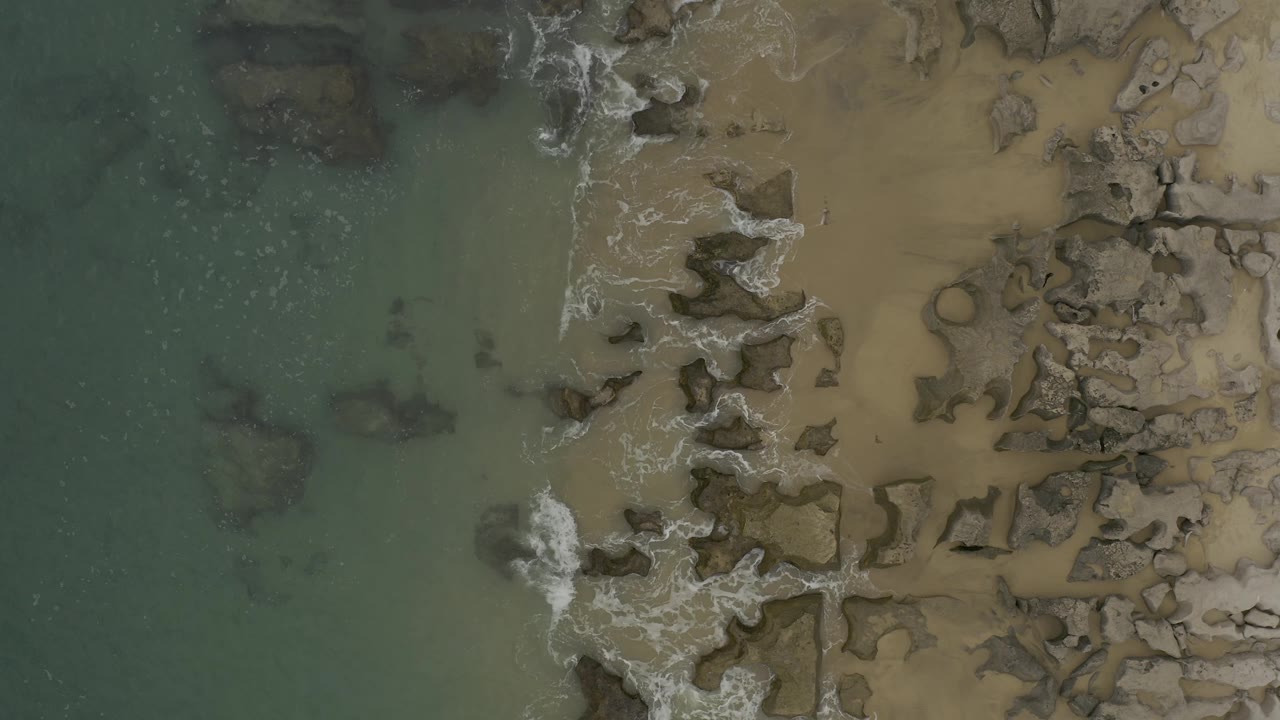 Overhead frame of Baja California Sur, Mexico, with green water left of frame, tan sand right of frame, and white surf spreading across clustered rocks with no visible people.