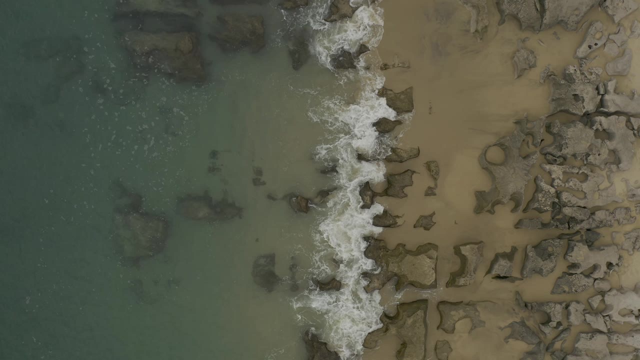 Aerial frame from Baja California Sur, Mexico, with white foam pushing farther across the central rock line, green water to the left, tan beach to the right, and no visible people.