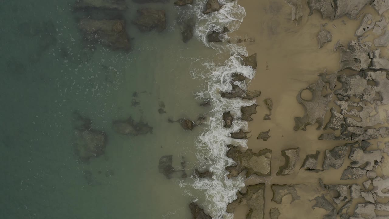 Top-down Baja California Sur shoreline view with a broken band of white surf, darker submerged rock shapes in the green water, tan sand on the right, and no visible people.