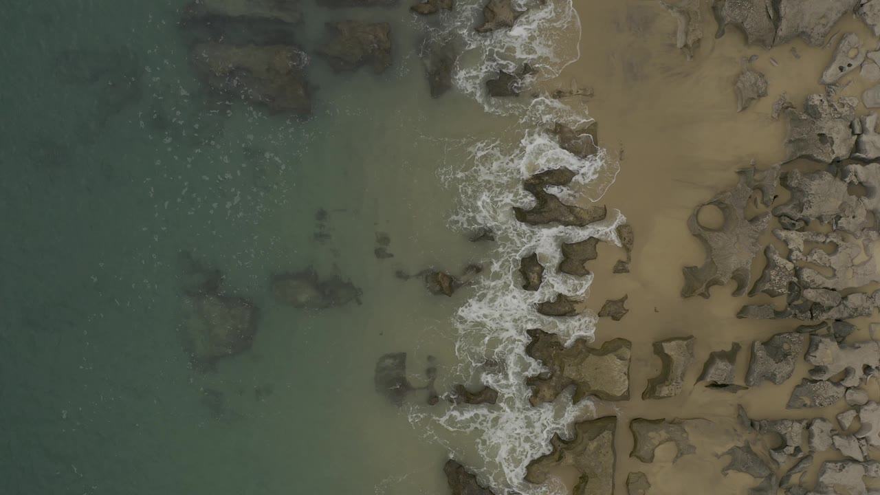 Overhead coastal frame in Baja California Sur, Mexico, where white waves break in a vertical strip between green water and a tan rocky beach, with no visible people.