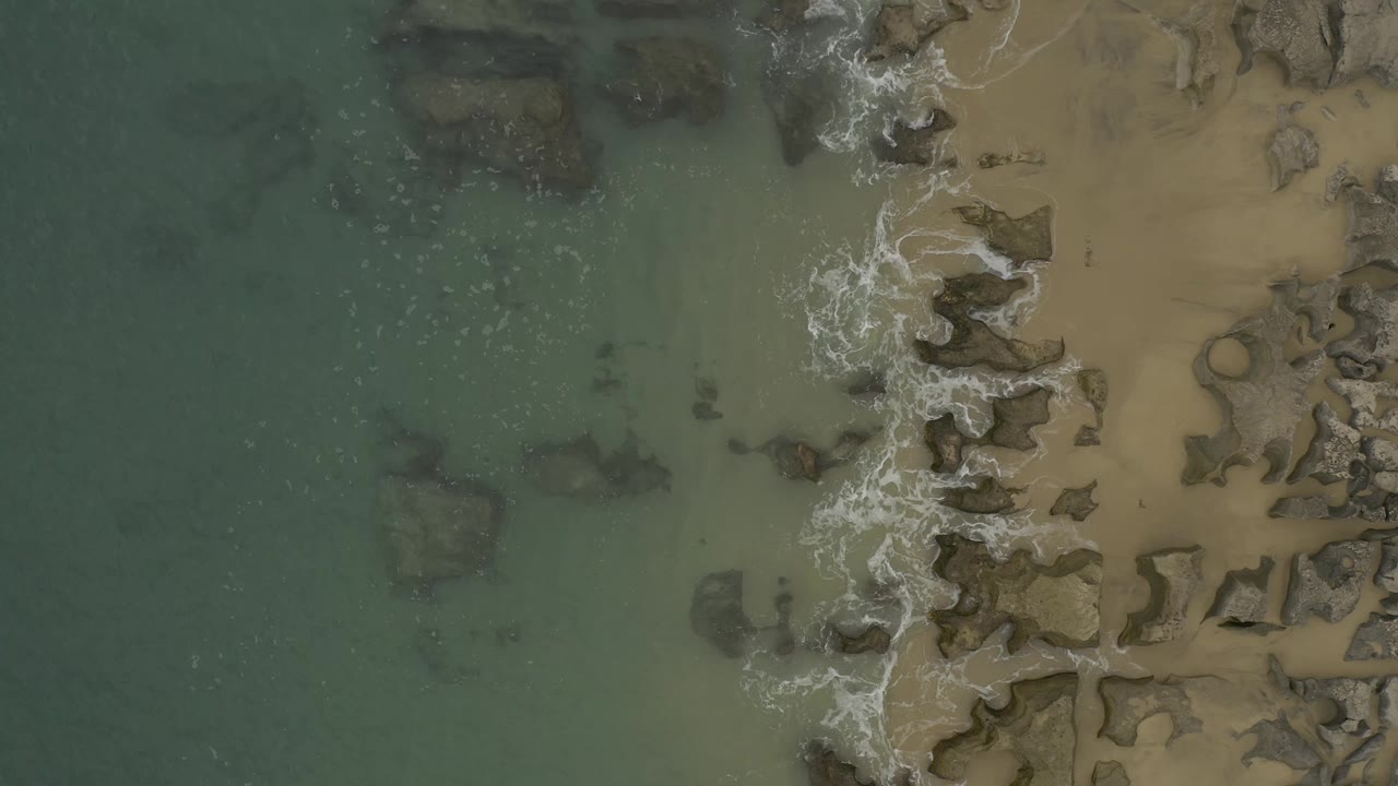 Aerial view in Baja California Sur, Mexico with white foam running along a jagged line of grey rocks between shallow water and sand, with no visible people.