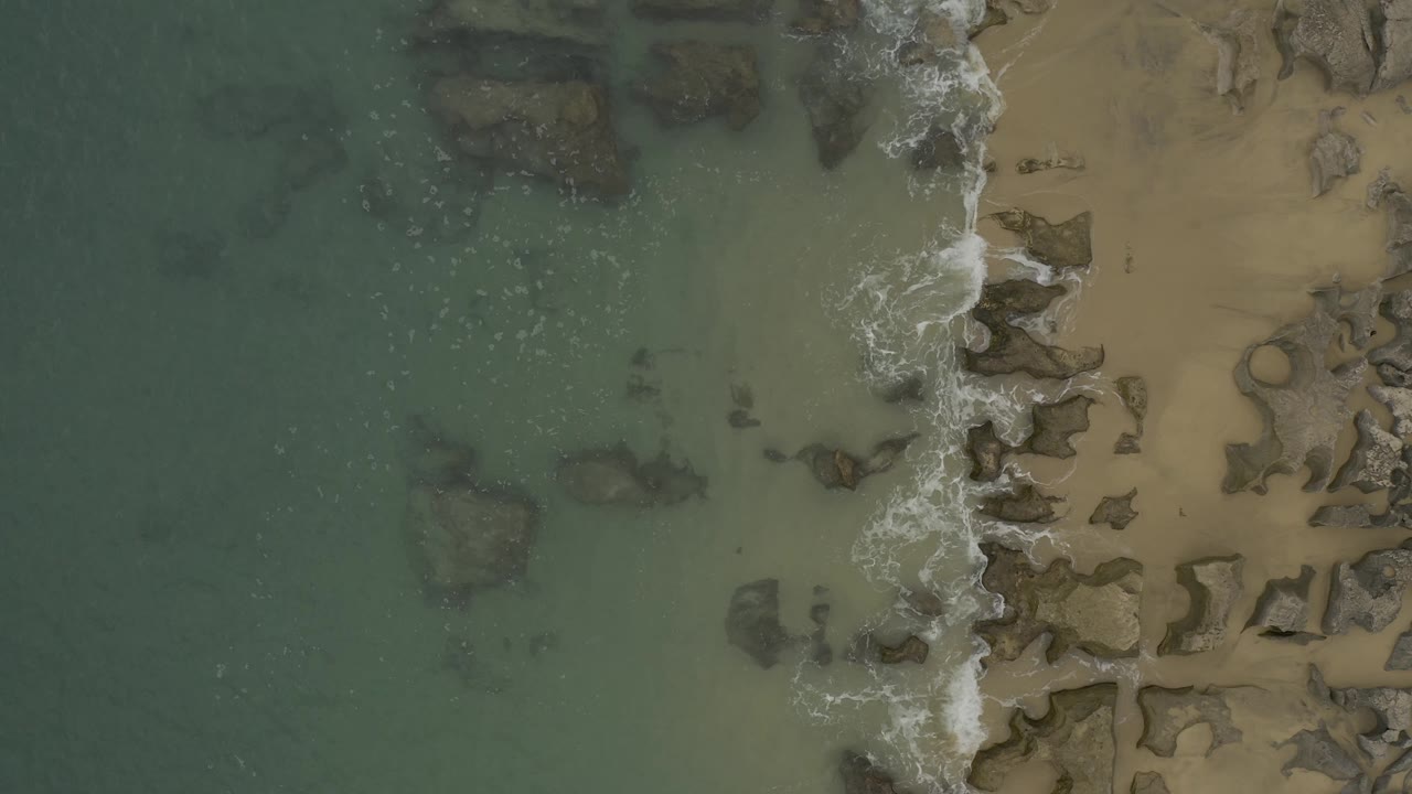 Top-down Baja California Sur coastline view with darker submerged rocks under blue-green water and a thin band of surf over the exposed rocks, with no visible people.