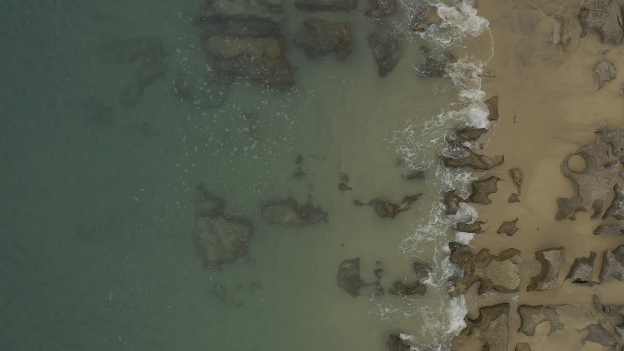 Overhead frame from Baja California Sur, Mexico showing clustered grey rocks along the shore, pale sand to the right, and green water to the left, with no visible people.