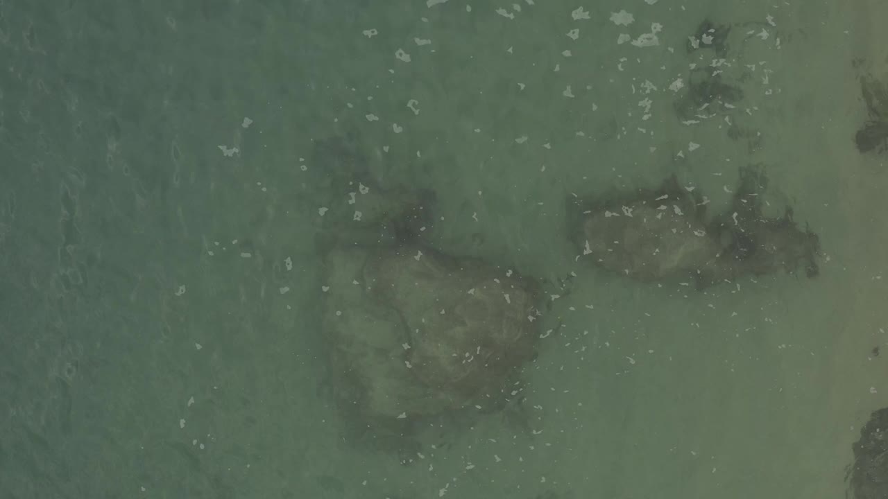 Aerial view over shallow green-blue water in Baja California Sur Mexico, with pale sand, dark submerged rocks, scattered foam flecks, and no visible people.