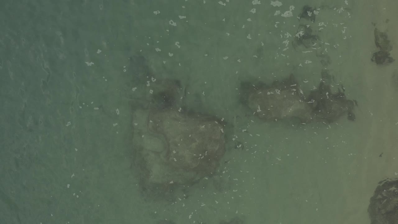Overhead Baja California Sur Mexico coastal water with a larger dark rock form near the lower area, a thin sandy strip at right, scattered white foam, and no visible people.