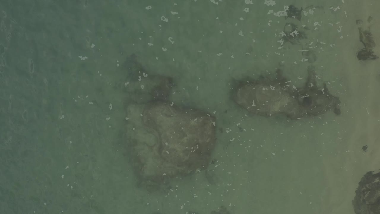 Top-down view in Baja California Sur Mexico showing pale sandy shallows, several dark submerged rocks, faint wave texture, and no visible people.