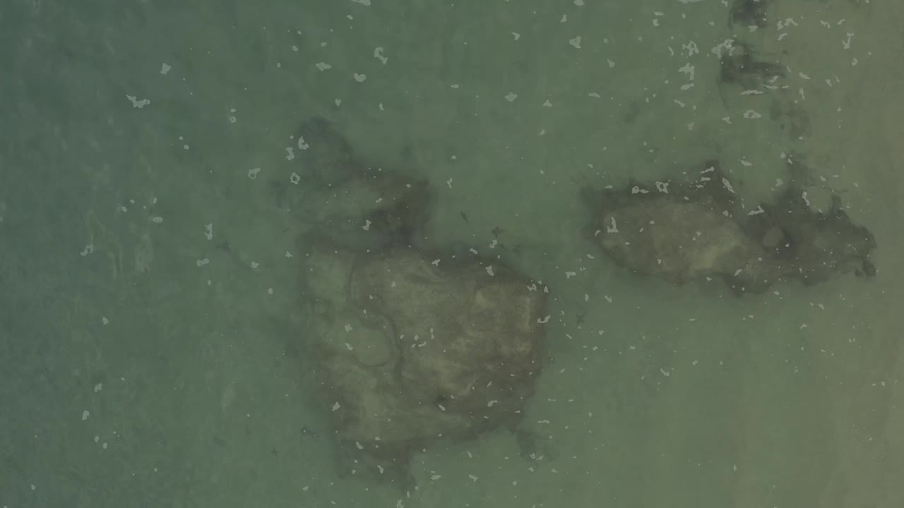 Aerial frame of Baja California Sur shallows with dark rock shapes under green-blue water and small white foam marks across the surface, no visible people.