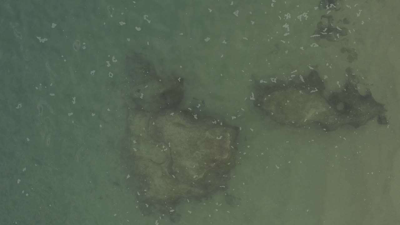 Overhead Baja California Sur coastal water shows submerged rocks through green shallows, with a paler sandy area along the right side and no visible people.