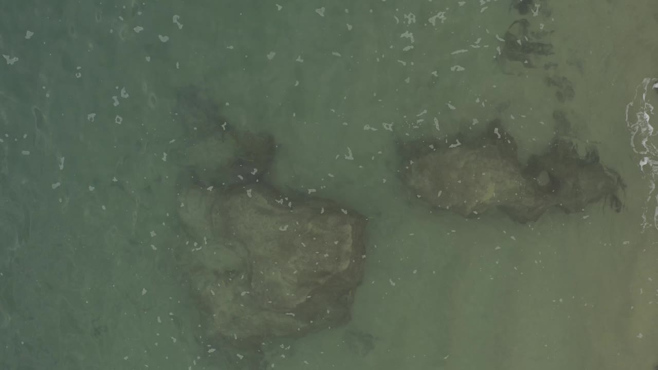 Baja California Sur overhead ocean frame with darker submerged rocks lower in the view, muted green water, and white foam gathering near the right edge, no visible people.