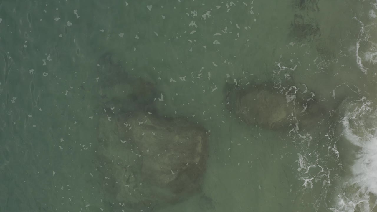 Top-down view of green-blue ocean in Baja California Sur, Mexico, with dark submerged rock shapes, scattered white foam, and no visible people.