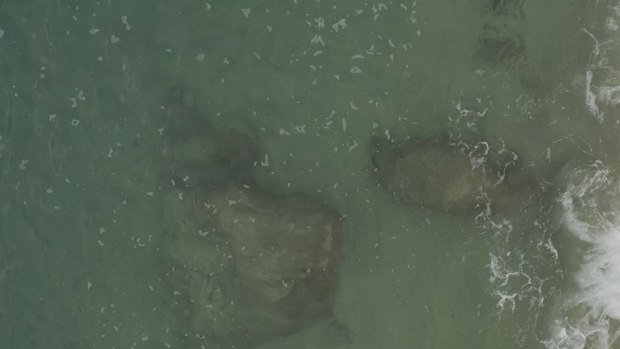 Overhead Baja California Sur ocean frame with muted green water, faint submerged forms, a brighter foam patch near the right side, and no visible people.