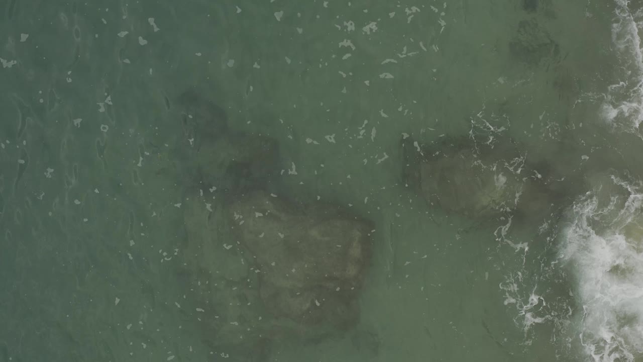 Top-down ocean view in Baja California Sur, Mexico, where dark underwater forms sit below green water and white foam gathers along the right edge, with no visible people.