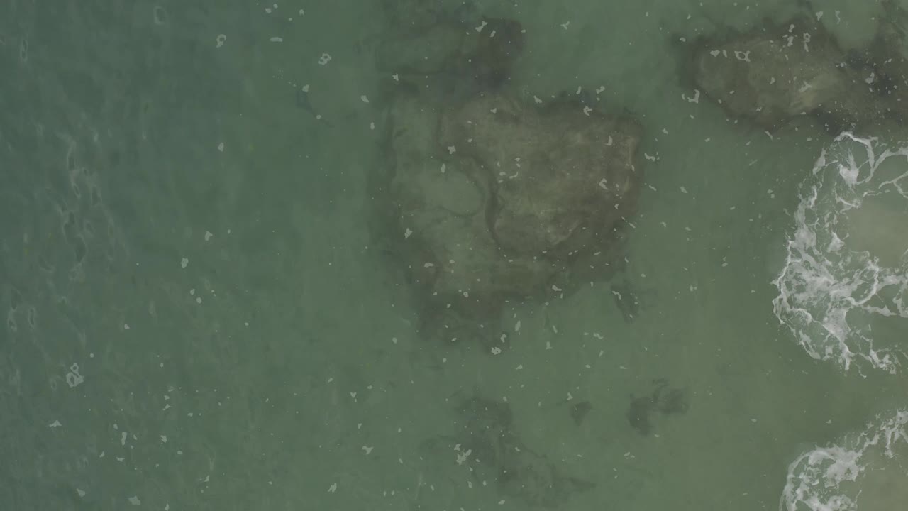 Aerial frame of shallow Baja California Sur ocean water where dark rocks sit below the surface and a white foam curl forms near the right edge, with no visible people.