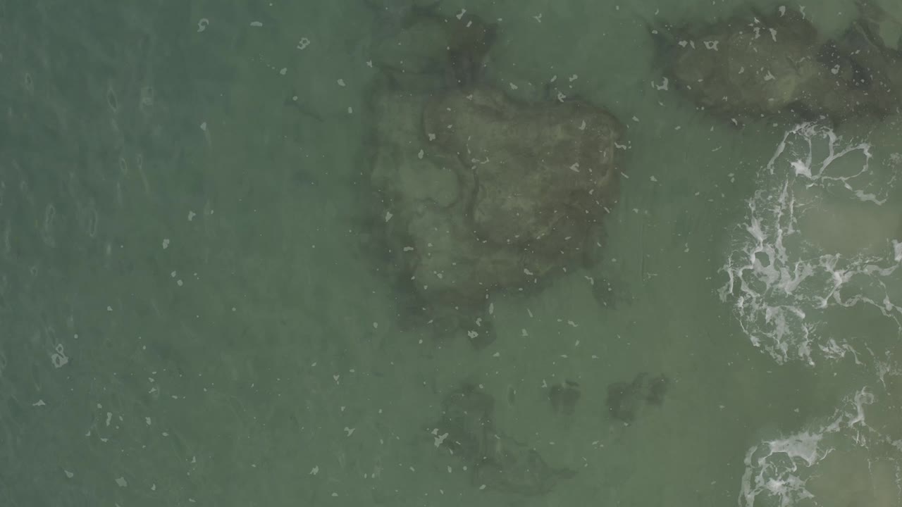 Top-down view of the Baja California Sur tidal zone with green water, shadowy rock shapes, and broken white foam markings across the right side, with no visible people.
