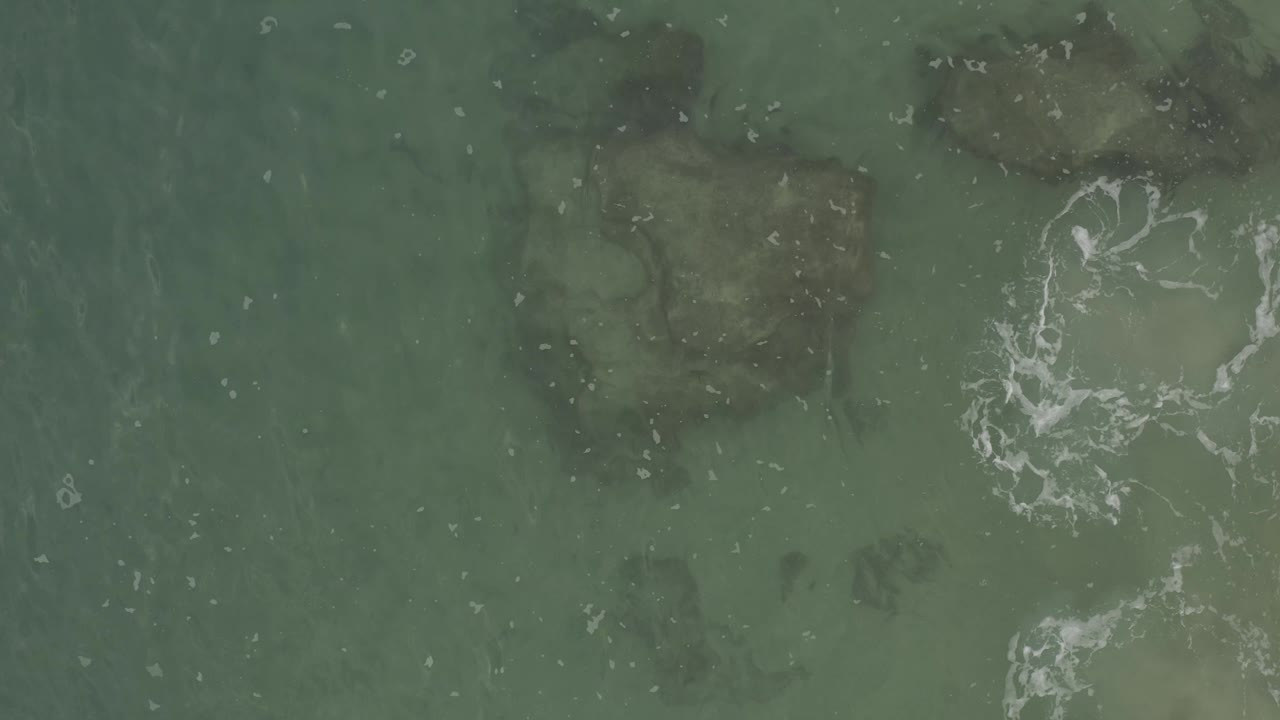 Overhead Baja California Sur ocean frame with muted green water, darker submerged rocks, and a loose cluster of white foam near center right, with no visible people.