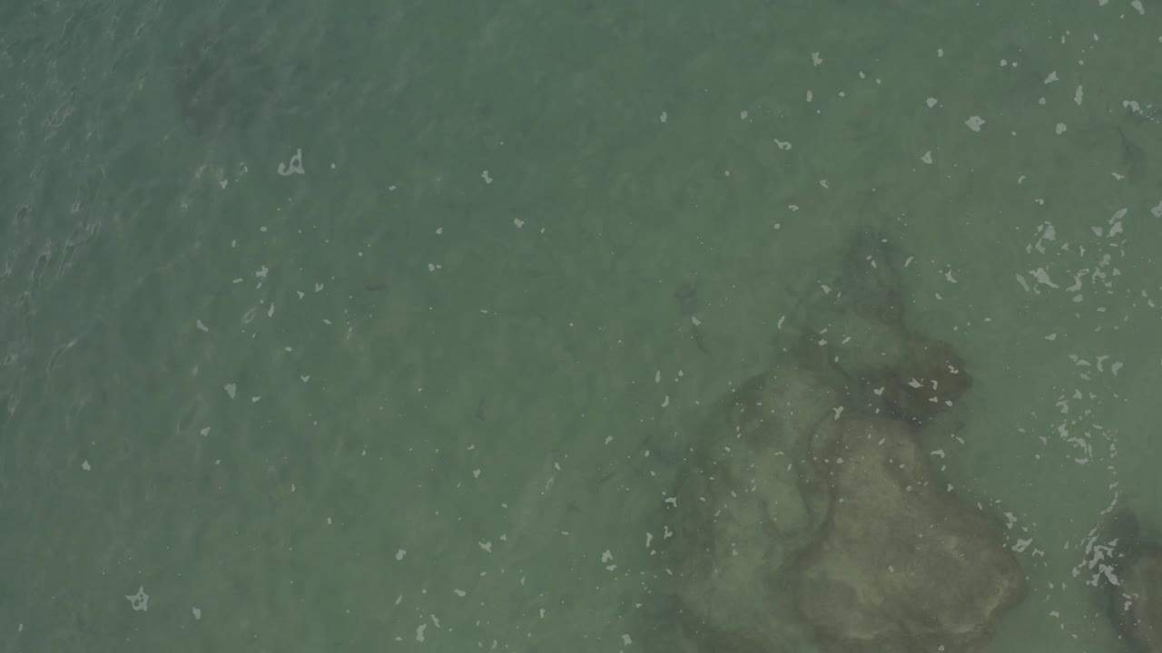 Top-down blue-green seawater with submerged rocks and white ripples.
