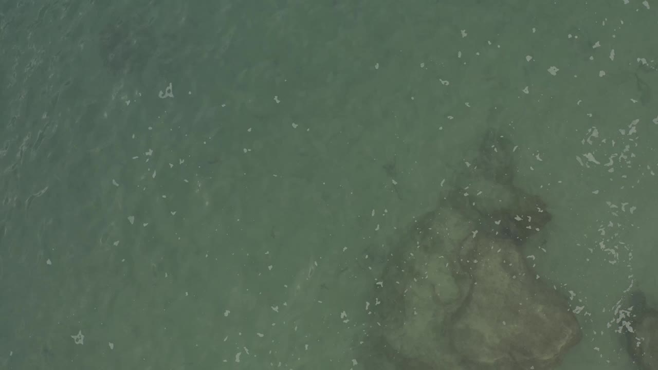 Aerial view of Baja California Sur seawater with a pale rock formation under the center of the frame, faint darker water patches, and no visible people.