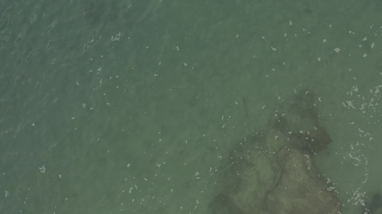 Clear blue-green water in Baja California Sur with submerged rocks sitting toward the right edge, scattered white foam flecks, and no visible people.