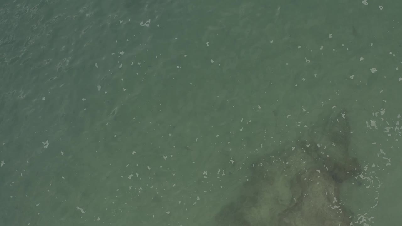 Top-down Baja California Sur ocean view with a muted rock shape low in the frame, translucent green water above it, white ripple marks, and no visible people.