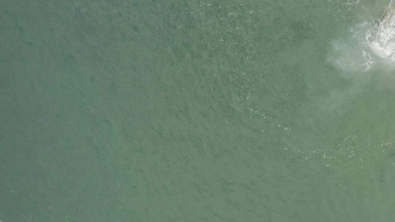 Aerial frame over Baja California Sur water, showing muted green ocean, subtle ripples, and a bright foam patch close to the top edge with no visible people.