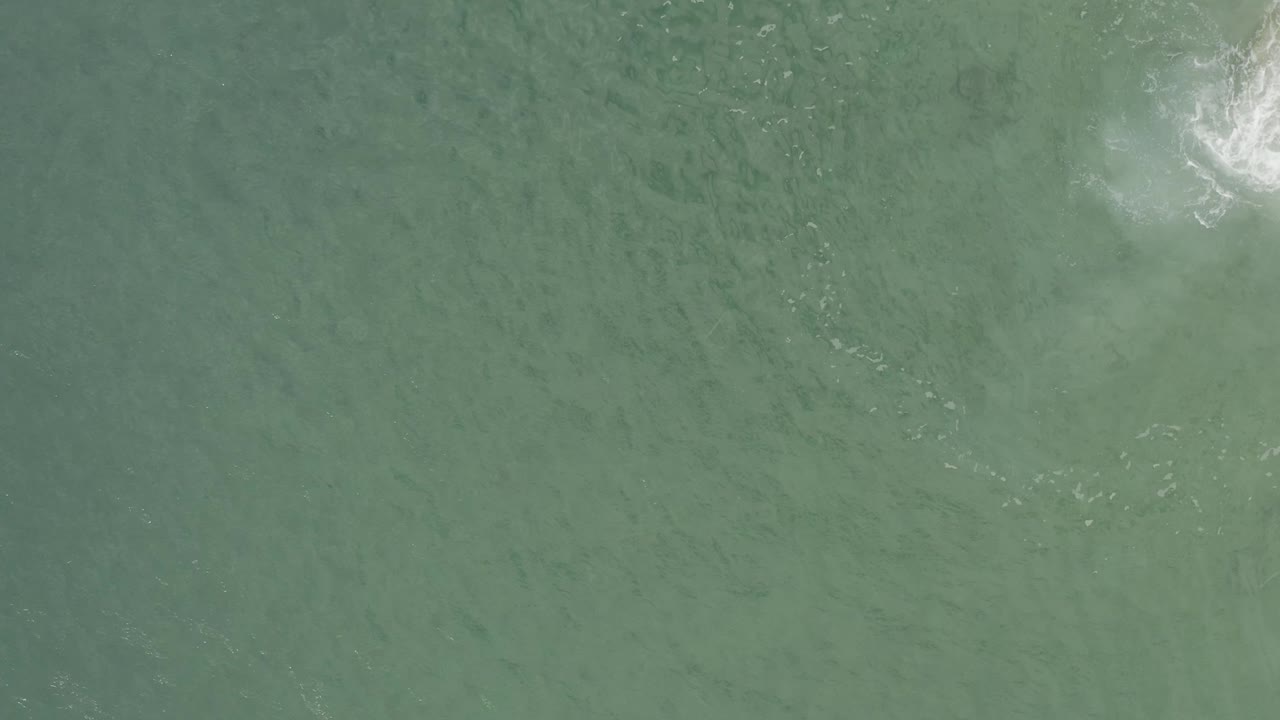 Straight-down aerial view in Baja California Sur, Mexico, with green water texture and white foam partly clipped by the upper right corner, no visible people.