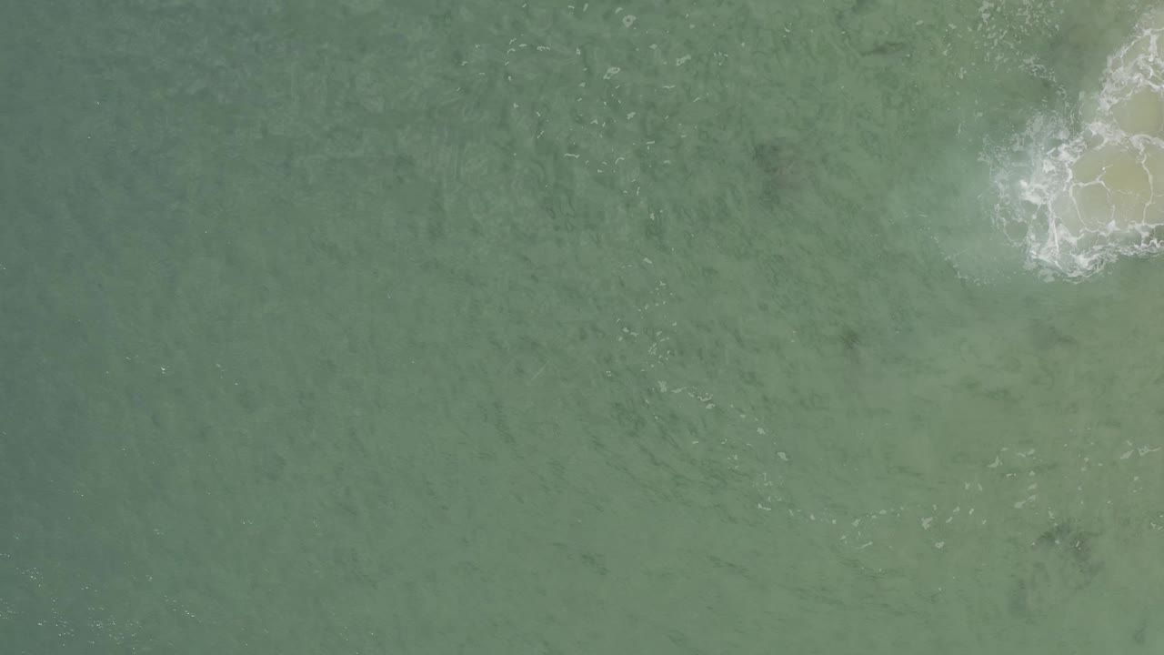 Top-down Baja California Sur ocean frame with muted green water and a foamy wave patch clipped by the upper right corner, with no visible people.