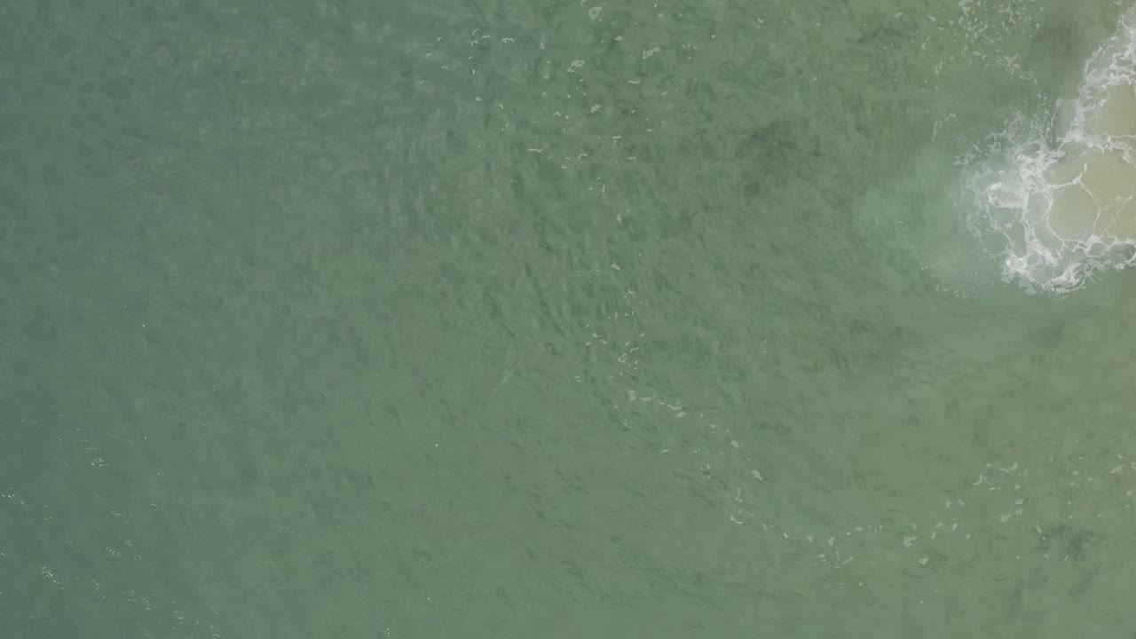 Overhead ocean view in Baja California Sur, Mexico, with white surf foam spreading farther along the right edge over a pale submerged patch and no visible people.