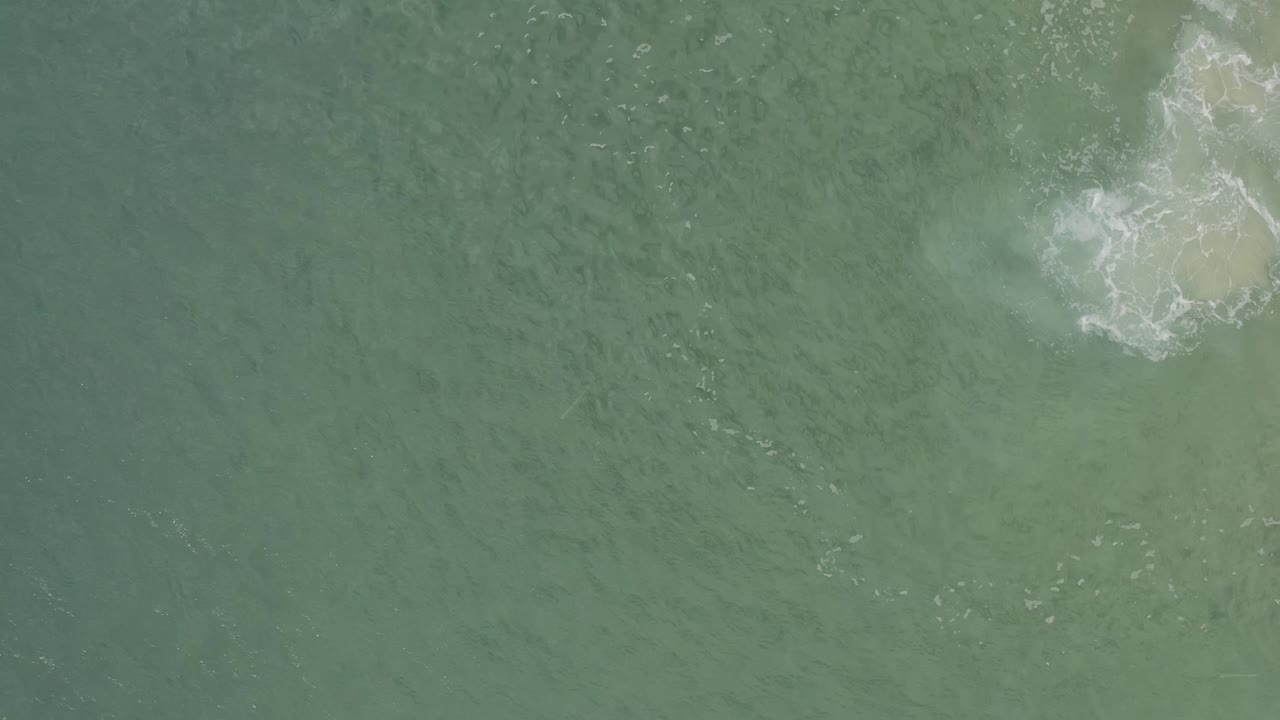 Aerial green water in Baja California Sur, Mexico, with white foam curling around a pale submerged area near the top right and no visible people.