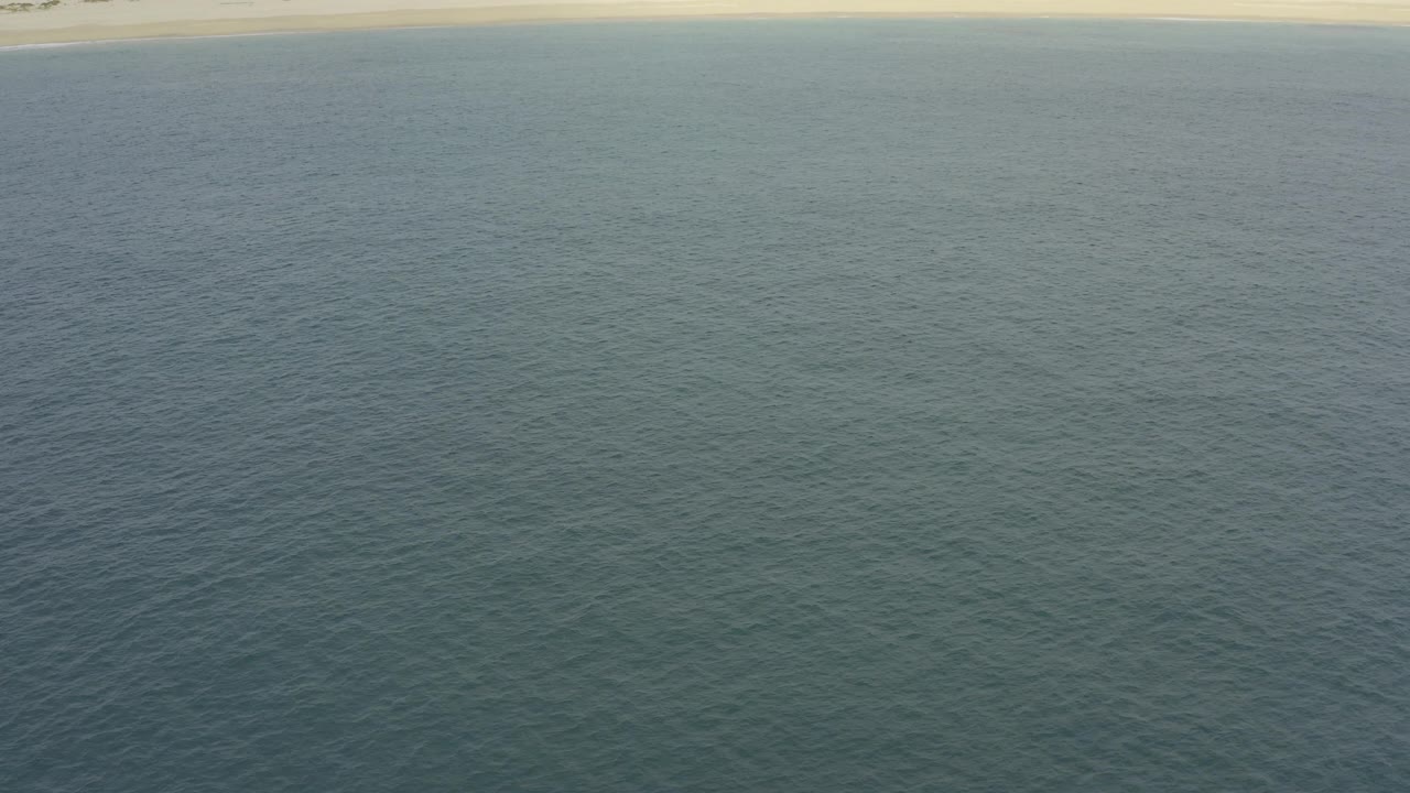 Wide elevated frame over textured water and a thin Baja California Sur sand beach, with low dunes beyond and no visible people.