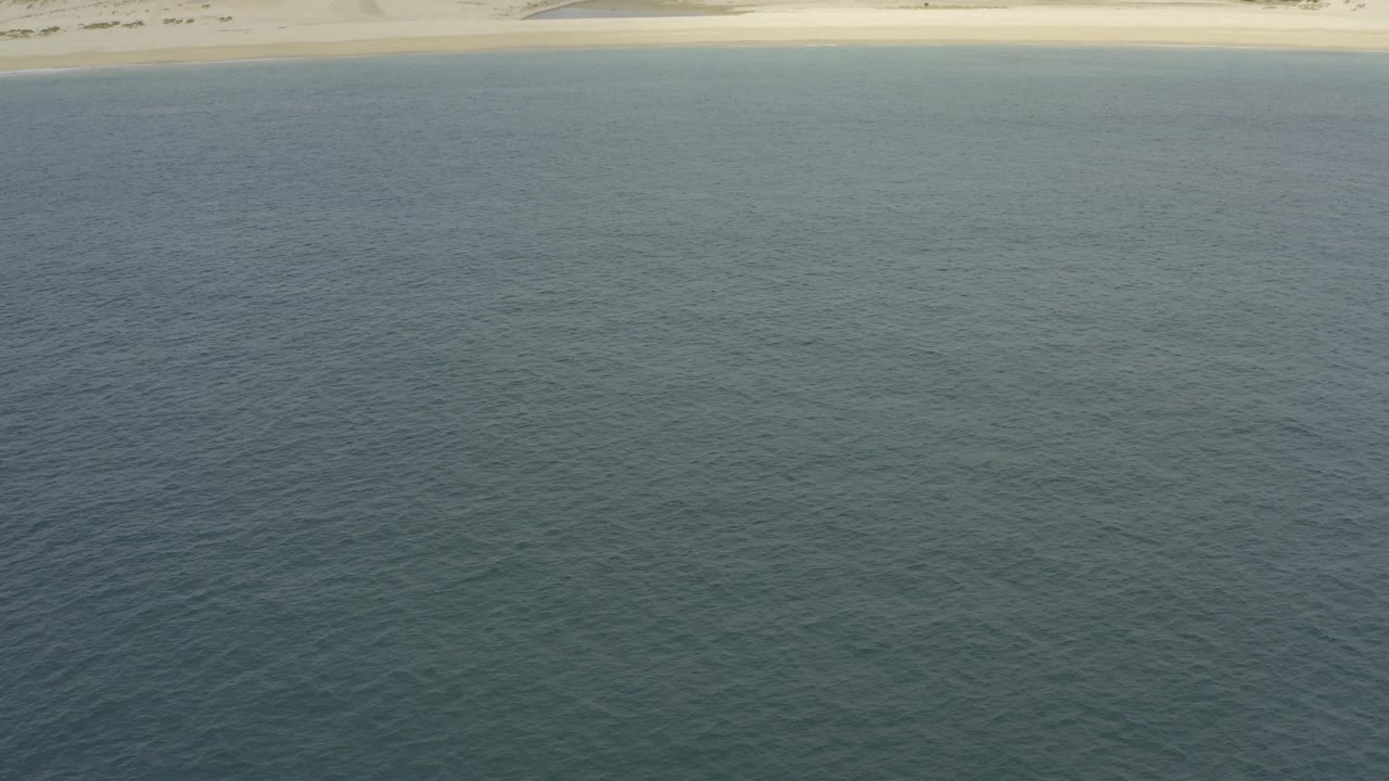 Elevated Baja California Sur shoreline view with rippled blue water in front, pale sand, dune shapes, scrubland, and no visible people.