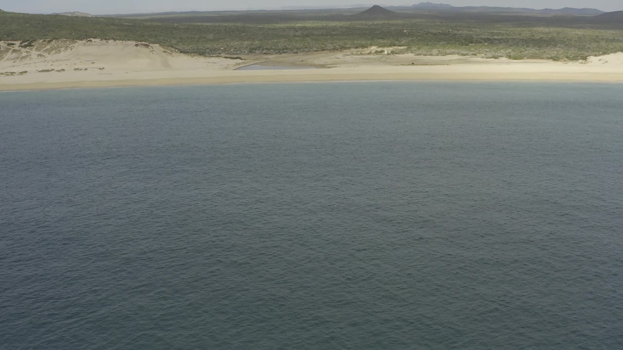 Aerial frame of the Baja California Sur shore with rippled blue water below, a pale tan beach band, dune ridges, low green scrubland, distant hills, and no visible people.