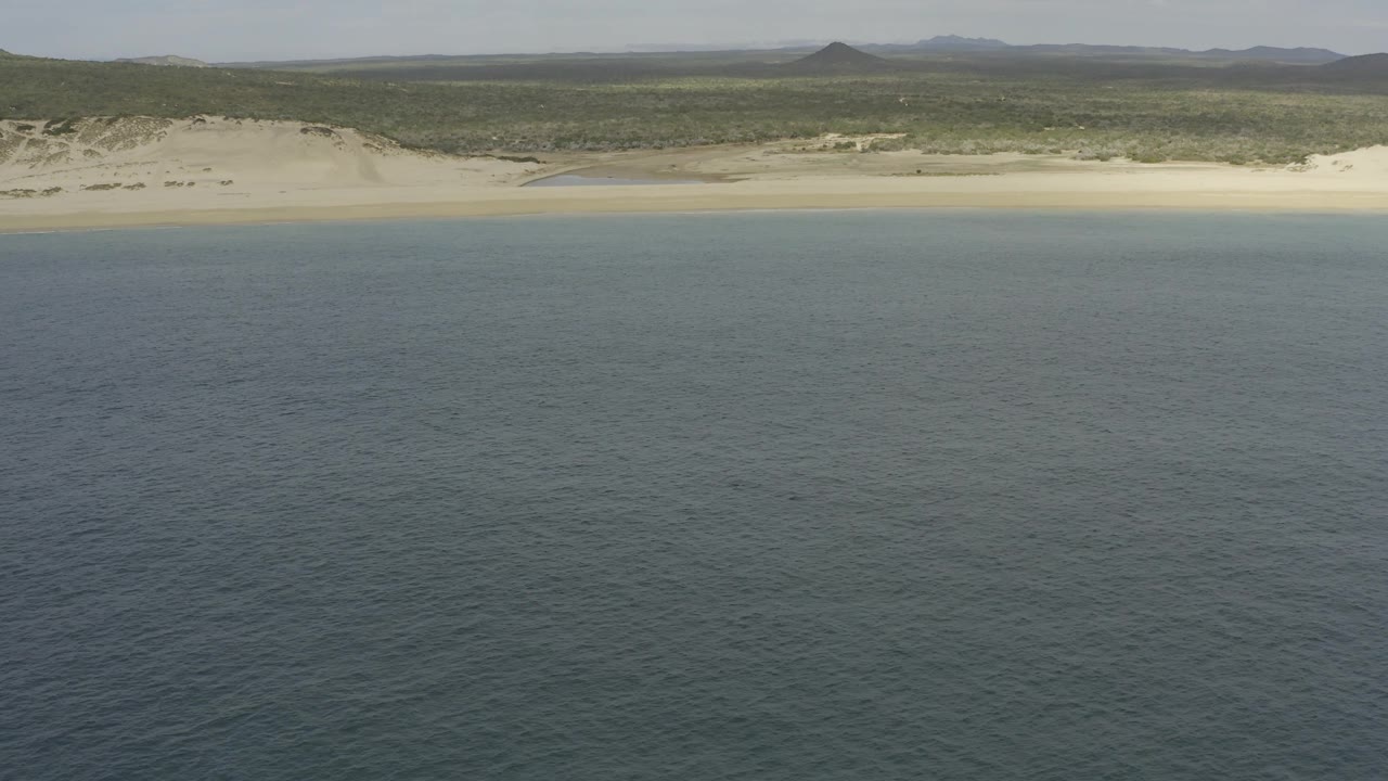 High aerial frame in Baja California Sur looking across dark blue ocean water toward a narrow tan beach, pale dunes, green inland scrub, low hills, and no visible people.