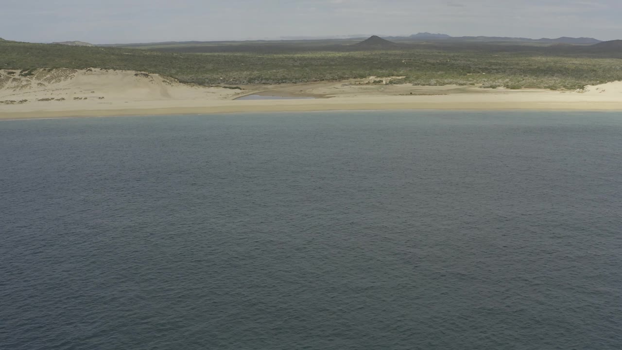 Blue ocean foreground with a long tan beach, dunes, scrubland, and distant hills under pale sky.