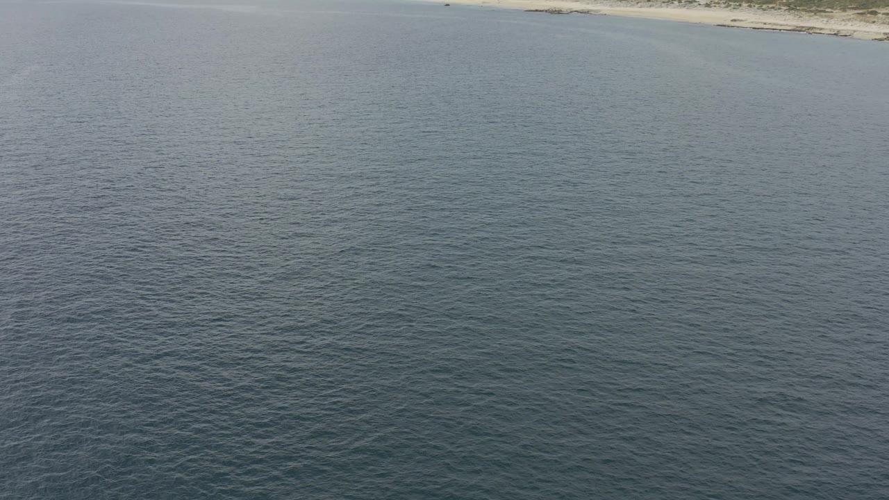 Aerial frame from Baja California Sur, Mexico showing blue water below, a tan beach across the upper frame, low green-brown hills beyond it, and no visible people.